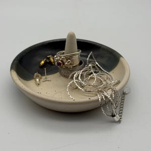 May include: A round ceramic ring dish with a two-tone black and cream speckled glaze. A central cone holds two stacked silver rings, one featuring a small red stone. A silver chain necklace and two small stud earrings rest on the dish surface.