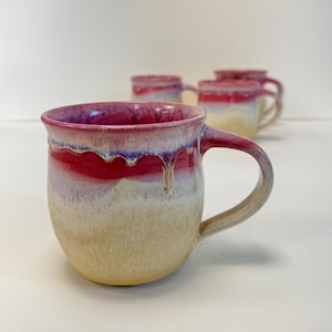 May include: A ceramic mug with a rounded body and a large handle. The mug has a gradient of colours, including a deep purple rim, a vibrant red band, and a creamy beige base. Several similar mugs are in the background.