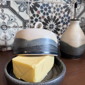 May include: A ceramic butter dish with a lid, in shades of blue and grey. It contains a block of yellow butter. An oil bottle is also visible. Kitchen accessory.