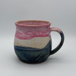 May include: A ceramic mug with a rounded body and a large handle. The mug has a colour gradient, with a pink rim, a white and beige middle section, and a dark blue and green base. The handle is dark blue.