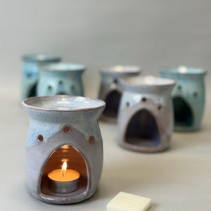 May include: A collection of ceramic oil warmers in pastel shades of blue and lilac, each with a lit tea light. The warmers have a rounded shape with an arched opening and small holes. A white wax melt is placed next to the front warmer.