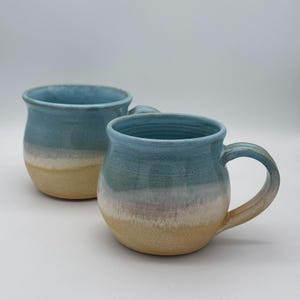May include: Two handmade ceramic mugs with a rounded shape and a handle. The mugs feature a gradient of colours, transitioning from a light blue at the top to a beige at the bottom. The mugs are suitable for coffee, tea, or other beverages.