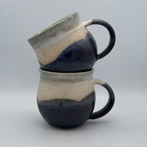 May include: Two stacked ceramic mugs featuring a rounded shape and a handle on the right side. The mugs have a tri-colour glaze pattern with a dark navy blue base, a central band of speckled beige, and a light sage green rim at the top opening.
