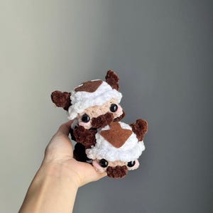 May include: Two handmade crochet plush toys, each resembling a brown and white creature with large black eyes and small pink accents. The toys are stacked on top of each other, showcasing the detailed craftsmanship and soft texture.