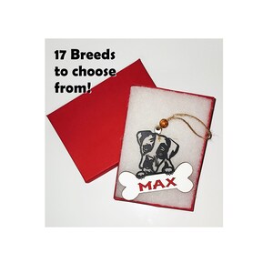 May include: A white wooden bone-shaped ornament with a black and white dog silhouette and the name "MAX" in red letters. The ornament is hanging from a red box with white foam padding.