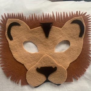 May include: A brown felt lion mask with black felt details. The mask has eye holes and a felt mane.