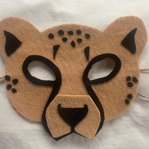 Könnte beinhalten: Eine braune Filz-Gepardenmaske mit schwarzen Augenlöchern und Flecken. Die Maske hat eine schwarze Filznase und Ohren. Die Maske ist mit einer Schnur gebunden.