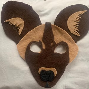 May include: A brown and tan felt mask of an African wild dog with black eyes and a black nose.