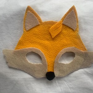 Op de afbeelding: Een vossenmasker van vilt met oranje vacht, bruine ogen en een zwarte neus. Het masker heeft twee grote, beige oren.