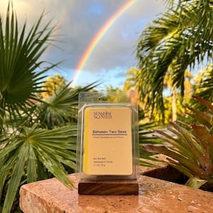 May include: A yellow soy wax melt in a clear plastic clamshell container with a label reading Sunshine Scented, Between Two Seas, Floral, Sandalwood and Ozone, Soy Wax Melt, Handmade in Florida, 2.7 oz / 76 g, resting on a small wooden base.
