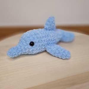 Puede incluir: Un juguete de delfín de felpa azul claro con un ojo negro. El animal de peluche hecho a mano tiene una apariencia texturizada y borrosa. El delfín está acostado sobre una superficie de madera clara. El juguete tiene una aleta en la espalda y dos aletas laterales.