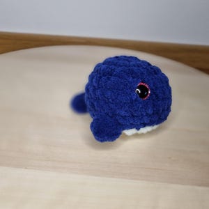 Könnte beinhalten: Ein handgemachter, plüschiger blauer Wal mit weißem Bauch und rosa umrandeten Augen. Der Wal ist aus einem weichen, strukturierten Garn gefertigt und hat eine kleine Schwanzflosse. Dieser entzückende Häkelwal ist ein süßes Spielzeug oder eine Dekoration für das Kinderzimmer.