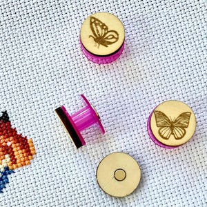 May include: Four circular needle minders featuring light wood tops with laser-engraved butterfly designs and bright pink translucent plastic bases. One needle minder is shown from the side, while the others display the butterfly motifs or a plain back.