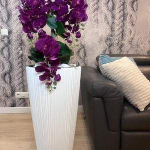 Könnte beinhalten: Ein hoher, weißer, geriffelter Pflanzkübel gefüllt mit leuchtend lila Orchideen. Die künstlichen Blumen fallen von der Oberseite des Pflanzkübels herab und erzeugen eine dramatische Darstellung. Der Pflanzkübel steht vor einem neutralen Hintergrund.
