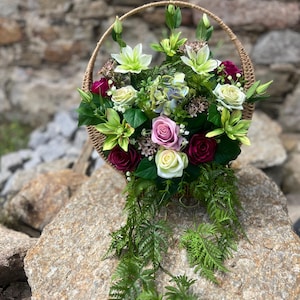 May include: A round woven basket filled with an arrangement of artificial flowers including white, pink, and deep red roses, light green lilies, and blue-tinted hydrangeas, accented with green fern fronds that drape downward from the basket base.