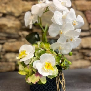 以下が含まれることがあります： 黄色い中心部と緑の葉を持つ白い蘭の花を使った装飾的なフラワーアレンジメント。 花は、テクスチャード加工された石の背景に対して、根が見える濃い青色の模様の正方形の容器に配置されています。
