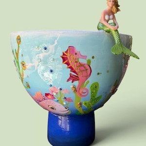 May include: A hand-painted ceramic bowl with a blue and turquoise underwater scene. The bowl features a pink seahorse, fish, and a mermaid figurine. The base is a solid blue color, and the bowl is decorated with aquatic plants.