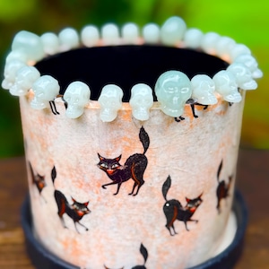May include: A cylindrical planter featuring a light orange and white mottled surface decorated with black cat illustrations. The rim is adorned with a continuous row of small, pale, translucent skull-shaped beads. The planter sits on a black base.