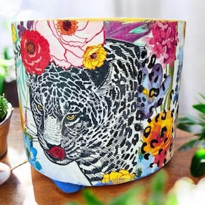 May include: A cylindrical planter pot featuring a black and white leopard design with colorful floral accents. The pot has a yellow rim and a blue base, creating a vibrant and artistic look. Perfect for indoor or outdoor plants.