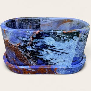 May include: An oval-shaped planter with a matching saucer, featuring a marbled finish in shades of deep blue, light blue, black, and copper. The surface displays a fluid, swirling pattern of colors across the exterior of the container.