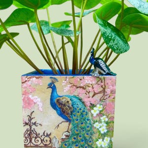 May include: A square planter featuring a painted peacock with blue and green plumage, standing near a black iron fence amidst pink and white flowers. A small, three-dimensional blue and green peacock figurine is attached to the top rim.