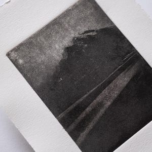 Puede incluir: Grabado monocromático de un paisaje, con una zona oscura y texturizada que sugiere una montaña o un acantilado. El grabado está enmarcado en papel blanco, creando un fuerte contraste. La obra es un estudio de luces y sombras.