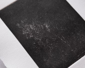 以下が含まれることがあります： テクスチャード加工された紙に印刷された白黒の抽象的なプリント。プリントは、より明るくテクスチャード加工された詳細を持つ暗い長方形の形状を特徴としています。紙にはデックルドエッジがあり、手作りの美しさを加えています。アートワークはミニマリストです。