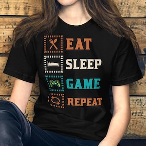 Könnte beinhalten: Ein schwarzes Kurzarm-T-Shirt mit einem Grafikdesign, das den Text EAT SLEEP GAME REPEAT in Orange, Weiß und Türkis zeigt. Jedes Wort wird von einem entsprechenden Symbol begleitet: Messer und Gabel, Bett, Gamecontroller und Pfeile.