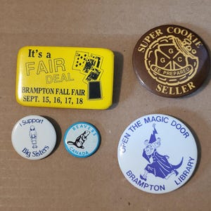 May include: A selection of vintage pin badges with diverse designs and text. A yellow badge says "It's a FAIR DEAL" with playing card symbols. Other badges feature "Super Cookie Seller," "Big Sisters," "Beavers Canada," and "Open the Magic Door."