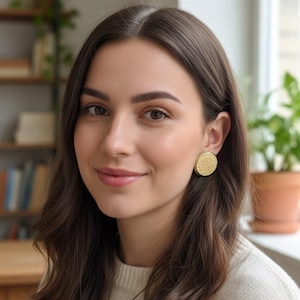 May include: A woman with long, wavy brown hair wearing a cream-coloured sweater and gold-coloured round earrings. The earrings have a textured surface. The background includes a bookshelf and potted plants.