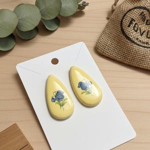 Puede incluir: Un par de pendientes amarillos en forma de lágrima con un diseño floral azul. Los pendientes se muestran en una tarjeta blanca. En el fondo, una pequeña bolsa de yute y una regla de madera.