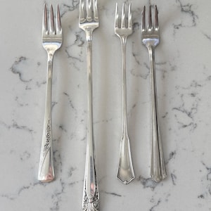 May include: Four silver forks with different handle designs are arranged on a marble surface. The forks vary in length and handle detailing, including ornate patterns and simple, streamlined styles. The tines are uniform in shape.