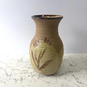 May include: A handcrafted ceramic vase with a textured, tan exterior and a dark brown rim. The vase features a light green base with brown leaf-like designs. The vase has a rounded body and a slightly flared top.