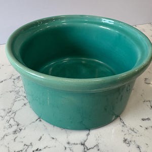 May include: A teal-colored ceramic bowl with a rounded shape and a slightly flared rim. The bowl has a smooth, glossy finish and appears to be empty. The bowl is sitting on a white and gray marble surface.