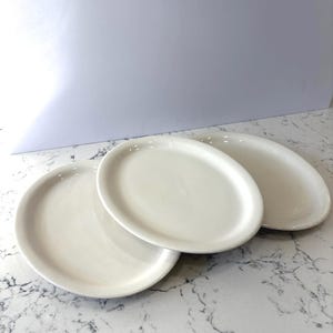 May include: Three off-white oval plates are arranged on a marble-patterned surface. The plates are stacked slightly, showcasing their simple, clean design. The plates are a neutral color, suitable for various dining settings.