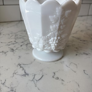 May include: A white, vintage-style milk glass bowl with a scalloped rim and a pedestal base. The bowl features a raised grape and leaf design on its sides, adding a touch of elegance. This decorative piece is perfect for serving or display.