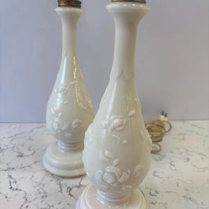 May include: Two off-white, vintage-style table lamps. Each lamp features a floral relief design and a brass-colored lamp holder. The lamps have a classic, elegant shape and are suitable for a bedroom or living room.