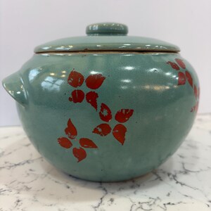 May include: A round, teal ceramic pot with a matching lid and two side handles. The pot is decorated with red floral designs. The pot is likely used for cooking or serving food.
