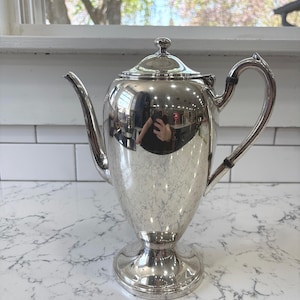 Puede incluir: Una cafetera plateada pulida con tapa, boquilla y asa curva. La cafetera se asienta sobre una base de pedestal. La superficie reflectante refleja el entorno. La cafetera se utiliza probablemente para servir bebidas calientes.