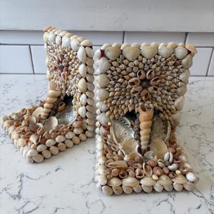 May include: Two decorative bookends crafted from various seashells. The bookends are rectangular with a curved top, and are covered in a variety of beige, tan, and white shells. The shells are arranged in a mosaic pattern, creating a textured surface.