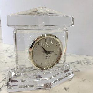 May include: A clear, crystal quartz clock with a silver-toned face and black hands. The clock is encased in a clear, geometric crystal structure with a rectangular base and a peaked top. The clock face displays the brand name "Lenox".