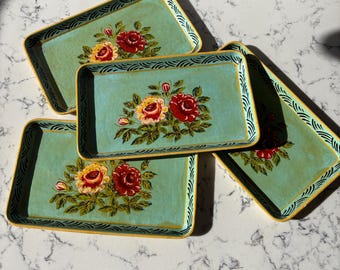 May include: Four rectangular serving trays with a light teal background and floral designs. The trays have a yellow border and a decorative black and white pattern. The floral design features red and yellow roses with green leaves.