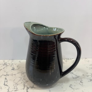May include: A dark brown ceramic pitcher with a curved handle. The pitcher has a textured, ribbed design and a light green interior. The rim is a lighter brown color. The pitcher is sitting on a white marble surface.