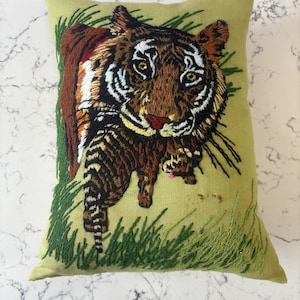 May include: Embroidered decorative pillow featuring a detailed tiger design. The tiger has brown, black, and white stripes, with green eyes and a pink nose. The pillow is rectangular, with a light green background and green embroidered grass.