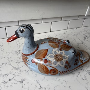 May include: A hand-painted ceramic duck figurine in shades of blue, brown, and red. The duck features floral and leaf designs on its body, with a red beak and a detailed eye. The figurine is a decorative item.