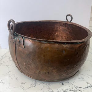 May include: A hammered copper bowl with two loop handles. The bowl has a rounded shape and a dark, aged patina. The copper has a rich, reddish-brown color with some areas of green oxidation. The bowl is likely an antique or vintage piece.