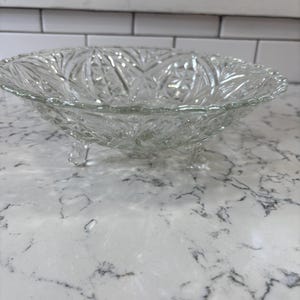 May include: A clear, decorative glass bowl with a detailed starburst and leaf pattern. The bowl has a scalloped rim and rests on three small feet. This vintage-style bowl is ideal for serving fruit or displaying decorative items.