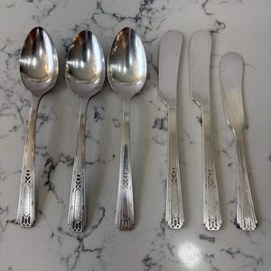 May include: A set of six silver-colored utensils, including three spoons and three butter knives, arranged on a marble-patterned surface. The utensils have a decorative geometric design on the handles.