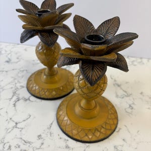 May include: Two decorative candle holders. The candle holders are gold-colored with a pineapple-shaped base and a flower-shaped top. The flower petals are brown. The candle holders are made of metal.