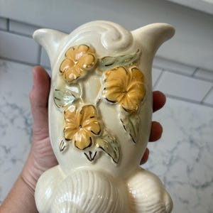 May include: Cream-colored ceramic vase with yellow floral accents and gold trim. The vase features a unique shape with a flared top and a textured base. The flowers are detailed with green leaves.